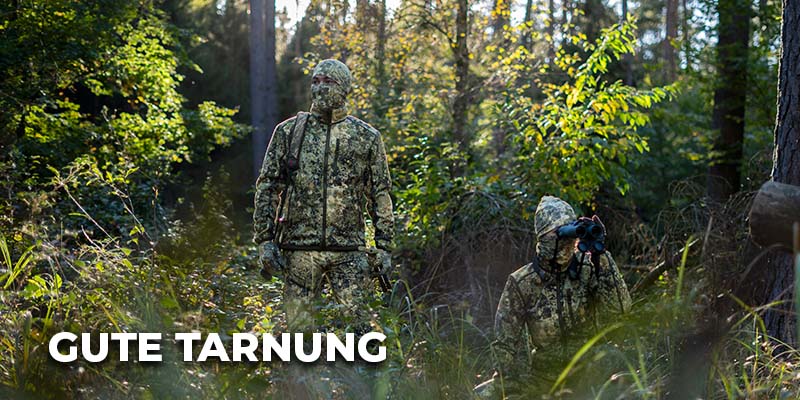 Zwei Jäger in vollständiger Tarnkleidung stehen und knien im dichten Unterholz eines Waldes; einer beobachtet die Umgebung mit einem Fernglas. Links unten steht der Schriftzug ‚Gute Tarnung‘.